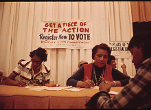 Three women are seated at a table while writing on a piece of paper with a sign hanging behind them that reads "Get a piece of the action - Register Now TO VOTE - Board of Election Commissioners - City of Chicago" 