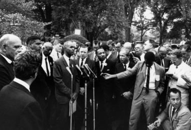 A group of organizers of the "March on Washington" stand together around microphones and a group of press
