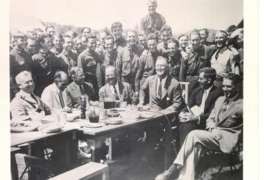 FDR having lunch while visiting Civilian Conservation Corps (CCC) camp, Co. 350, at Big Meadows, Shenandoah Valley, Virginia, August 12, 1933. Members of the CCC smile for the camera behind FDR and other seated officials.