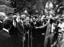 A group of organizers of the "March on Washington" stand together around microphones and a group of press